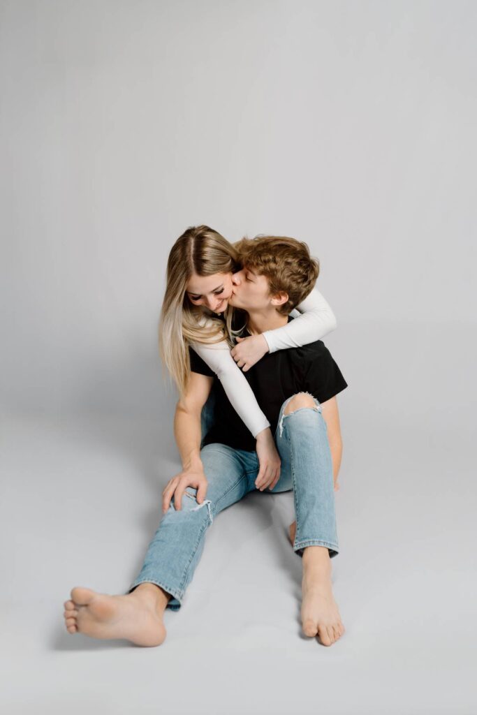 man sits on the ground and kissing his girlfriends cheek who has her arms over him and is sitting on a chair behind him