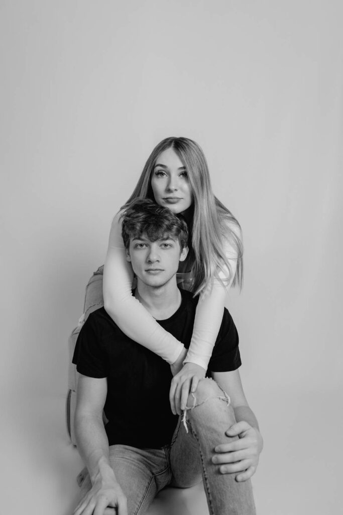 girl sitting on a chair with boy sitting down while her arms drape over his shoulders serious faces