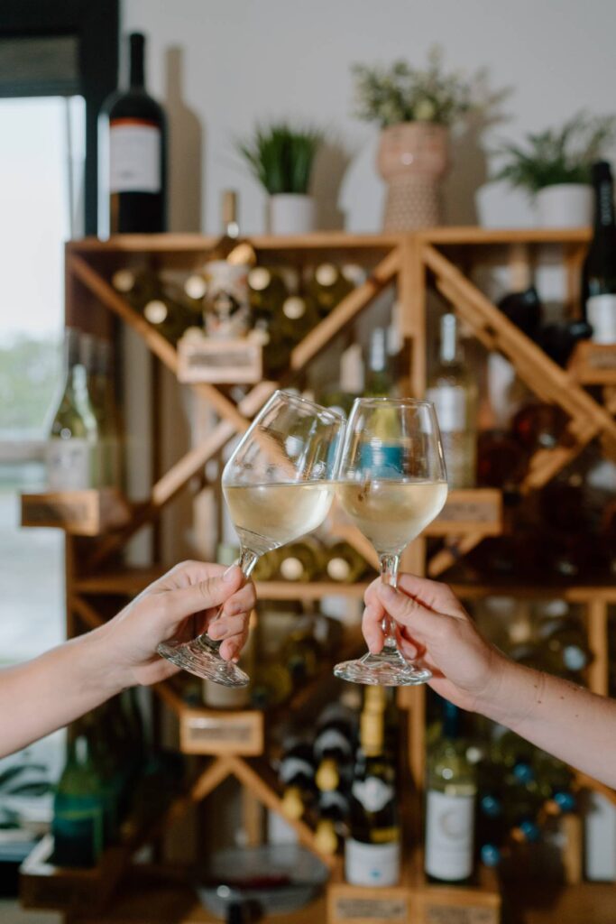 two people showing their hands holding two glasses of wine cheersing  in front of wine rack