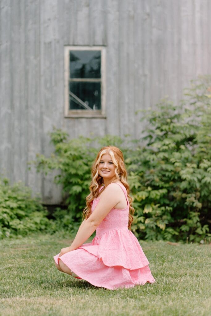 Senior girl in pink dress and cowboy boots standing by a rustic barn in Waupaca, WI

